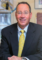 The image shows a man wearing glasses, with a beard, sitting at a desk with a microphone, smiling towards the camera.