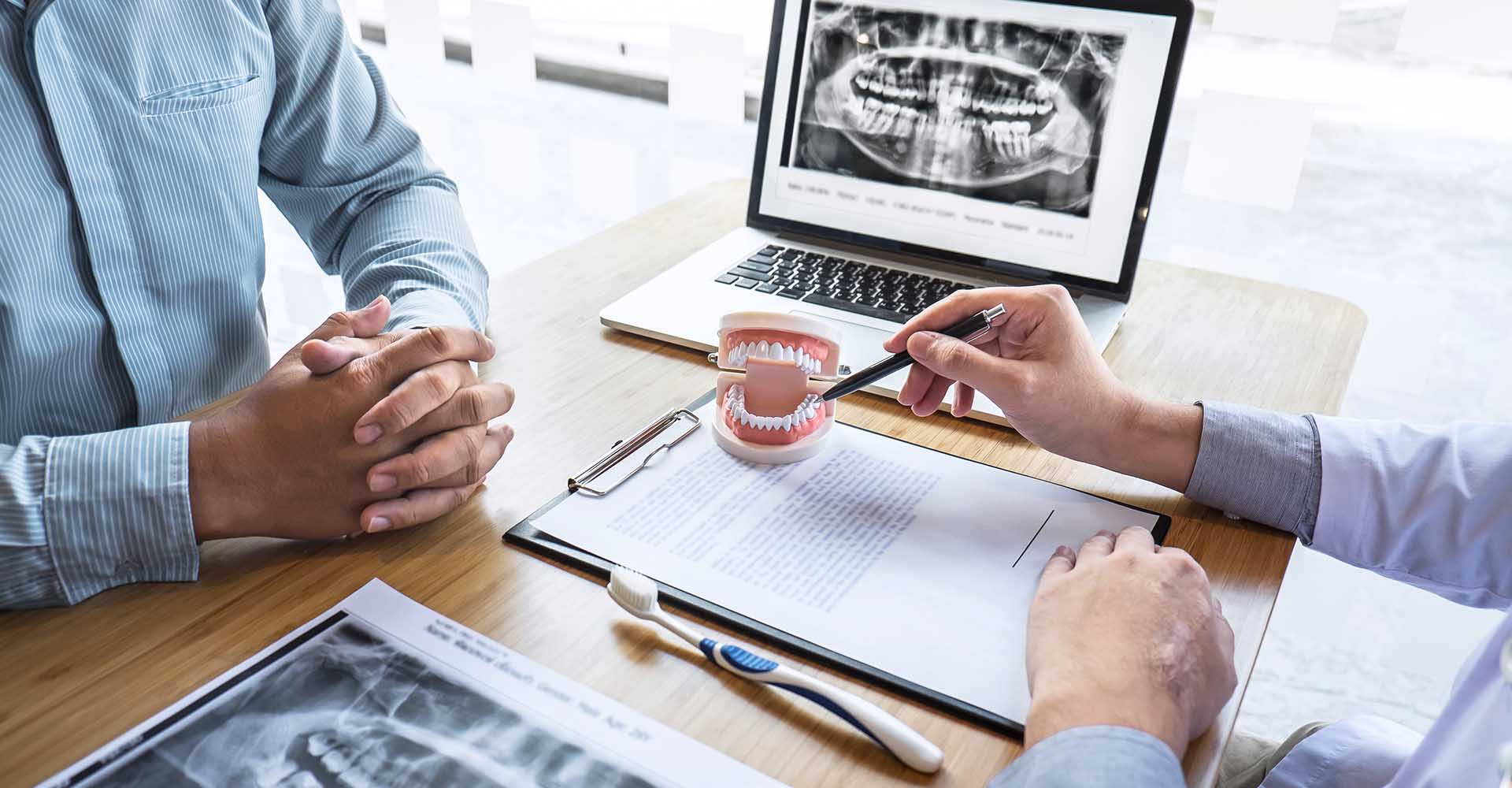 The image shows two individuals sitting at a table with a laptop displaying an X-ray image. A dental professional appears to be examining the X-ray, while another person is observing the screen. They are in a professional setting, possibly a dental office, with various documents and tools on the table.
